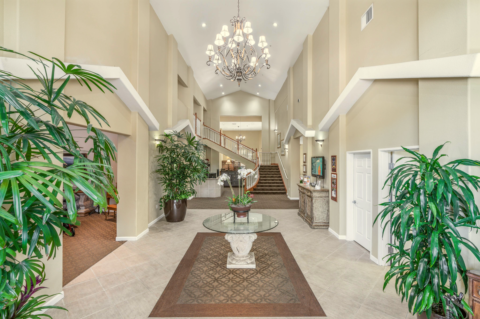 Spacious, elegant Eskaton lobby with high ceilings, a large chandelier, potted plants, a central round glass table, neutral walls, and a staircase leading to an upper floor in the background.