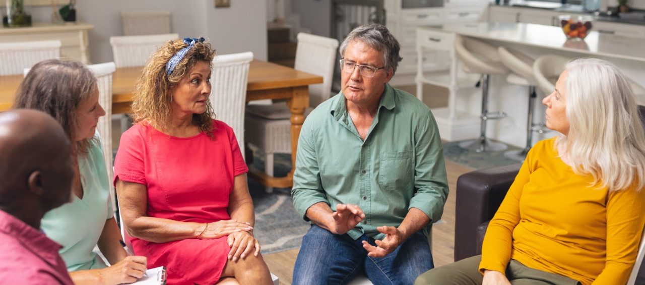 A group of five adults sits in a circle indoors, having a serious discussion as part of Eskaton support groups. They appear engaged and attentive, with one man in a green shirt talking while the others listen in a bright, modern room.
