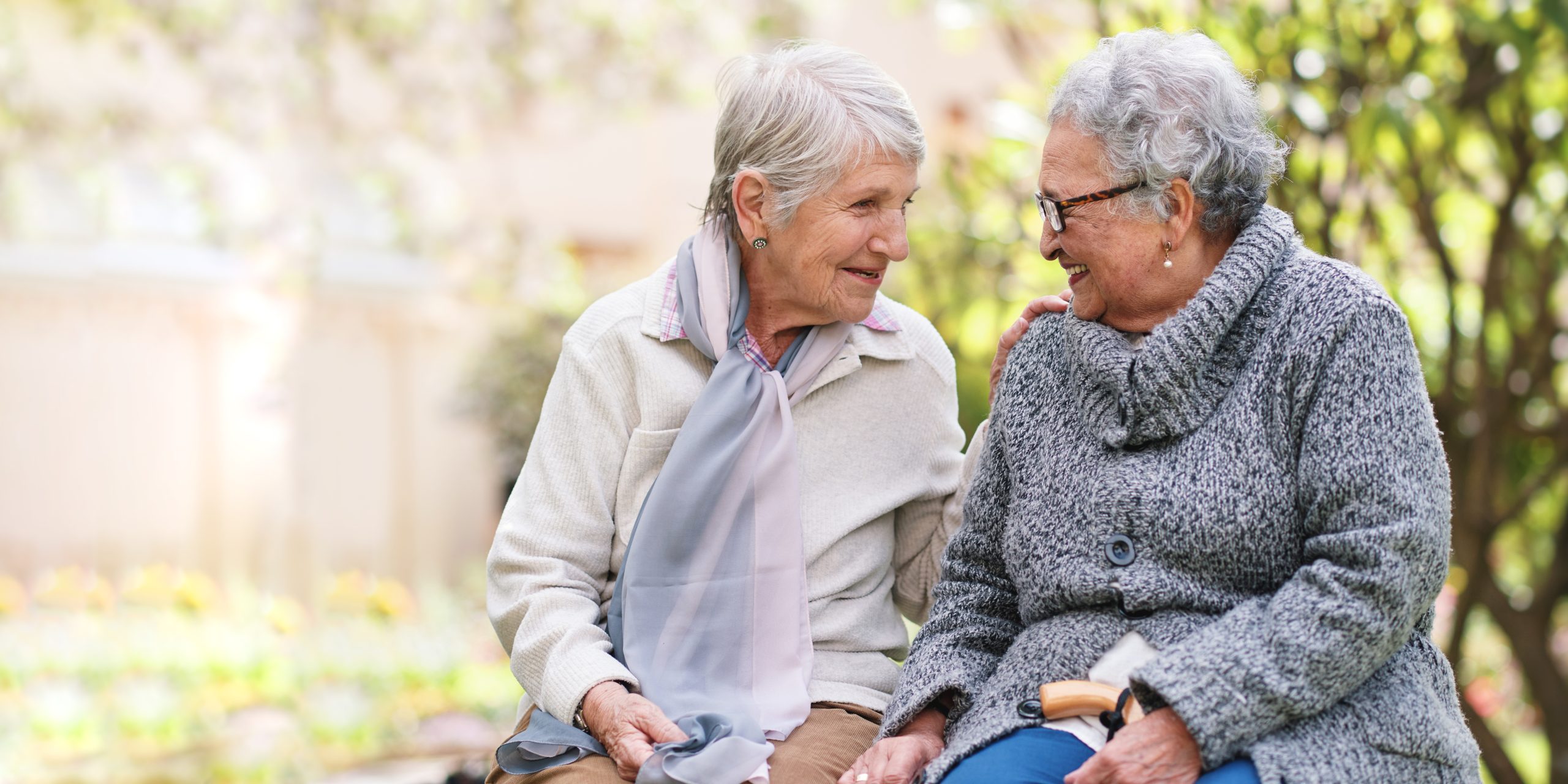 Two elderly women sitting on a bench outdoors, smiling and looking at each other warmly. They are dressed in cozy sweaters and scarves, surrounded by greenery and soft sunlight.