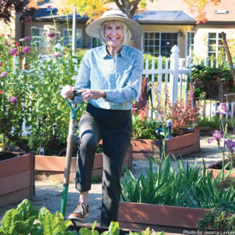 Smiling woman in a wide-brimmed hat stands in a garden, leaning on a shovel, surrounded by raised EVC Gardening beds filled with flowers and vegetables. A white picket fence and house are in the background.