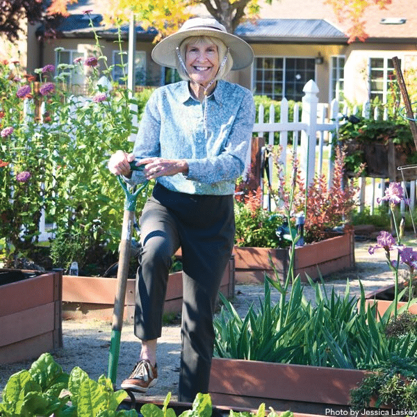 Smiling woman in a wide-brimmed hat stands in a garden, leaning on a shovel, surrounded by raised EVC Gardening beds filled with flowers and vegetables. A white picket fence and house are in the background.