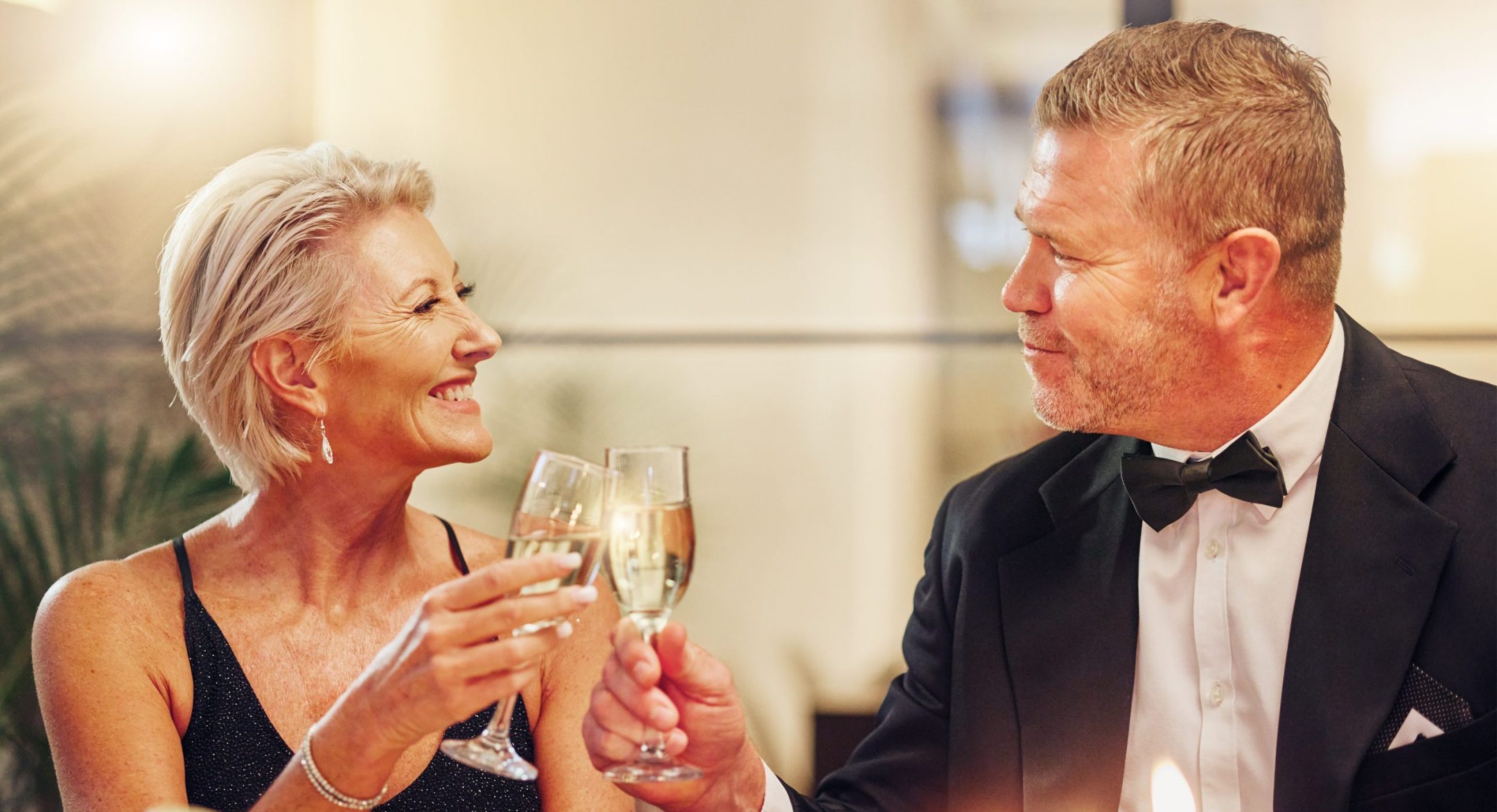 An older couple dressed elegantly smile and clink glasses of champagne at a candlelit dinner table, enjoying a celebratory or romantic evening together at the Longevity Gala.