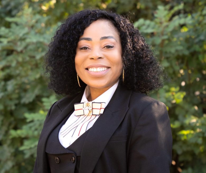 A woman with curly black hair, wearing a black suit and white shirt with a decorative bow, stands smiling in front of green leafy bushes at Eskaton senior living.
