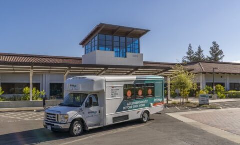 A white and green shuttle bus with Eskaton senior resources on the side is parked outside a modern building with large windows and a tile roof under a clear blue sky.