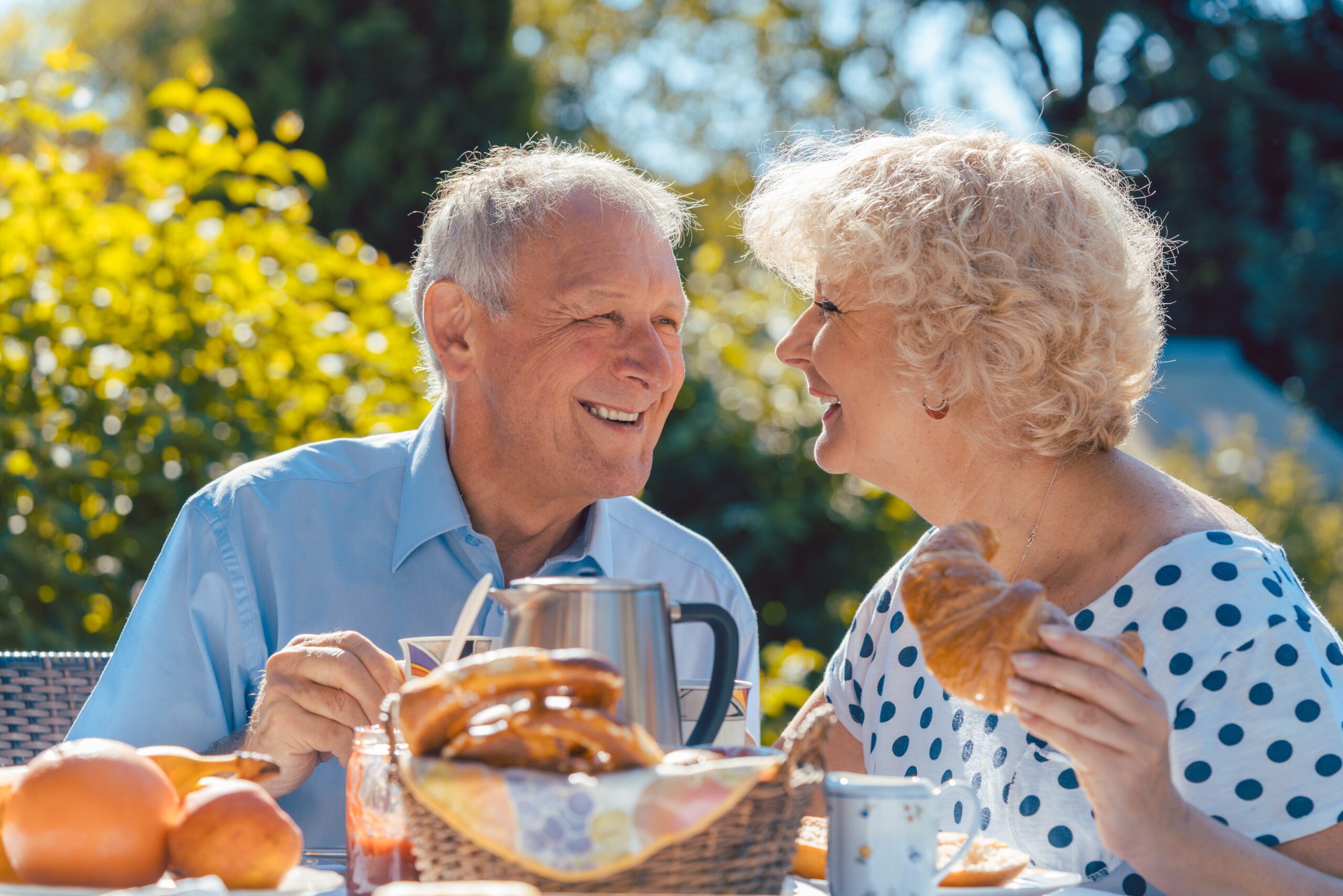 An elderly couple sits outdoors at a table, smiling and enjoying breakfast together. The woman holds a croissant, and there are pastries, fruit, and coffee on the table. Sunlight and greenery are visible in the background.