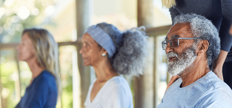 A group of older adults sit cross-legged with eyes closed, meditating peacefully in a sunlit room. The focus is on a man in front, attending one of the Eskaton support groups, with others sitting calmly behind him.