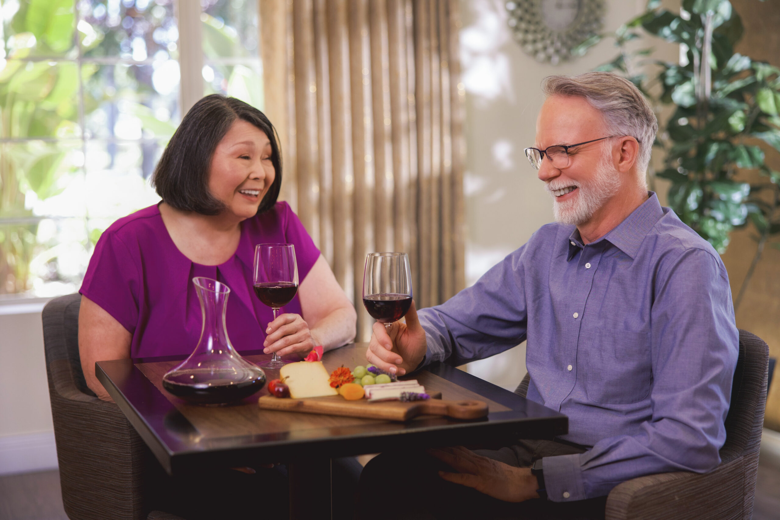 An older woman and man sit at a table, smiling and holding glasses of red wine. There is a decanter, assorted cheese, and fruit on the table. The setting appears bright and cozy with plants and curtains in the background.