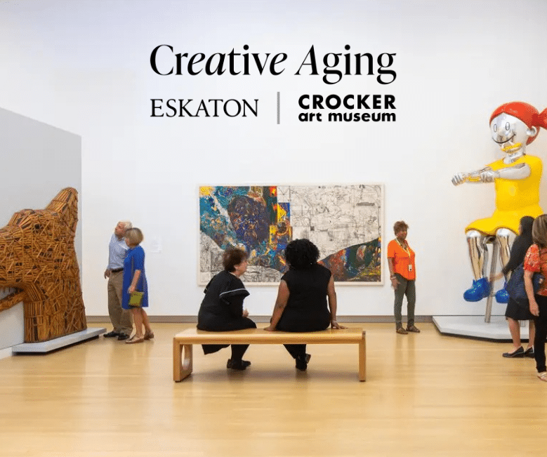 People observe colorful artwork and sculptures in a modern gallery. Two women sit on a bench, while others stand and look at the art. Creative Aging Eskaton | Crocker Art Museum text and Senior Care resources are displayed on the wall above.