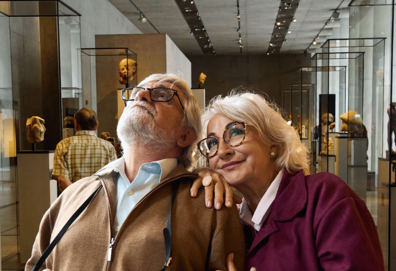 An elderly couple stands in a museum. The man, holding a camera, looks up at an exhibit, while the woman in a maroon coat smiles and leans affectionately on his shoulder—enjoying their visit thanks to Eskaton support groups. Glass cases and sculptures fill the background.