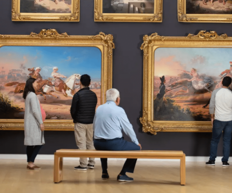 Several people observe large, ornate-framed paintings of dramatic horse scenes in an art gallery. As Eskaton support groups visit, one man sits on a bench while others stand and admire the artwork on the dark-colored wall.