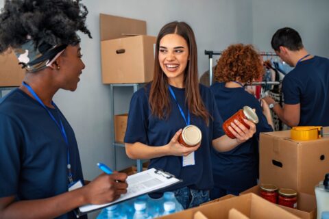 Four people wearing navy blue shirts work together in a room sorting canned and jarred food items. Two women smile and talk while holding jars; others pack boxes. Bottled water is visible on the table.