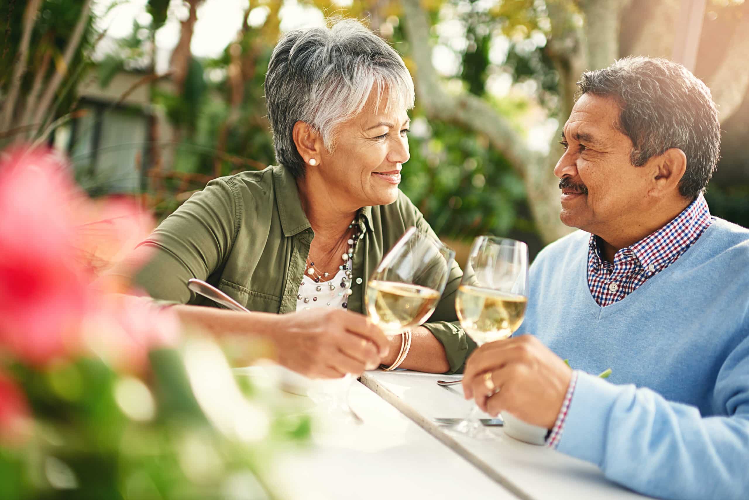 An older couple sits at an outdoor table, smiling at each other and holding glasses of white wine, enjoying a bright, sunny day with greenery and flowers in the background.