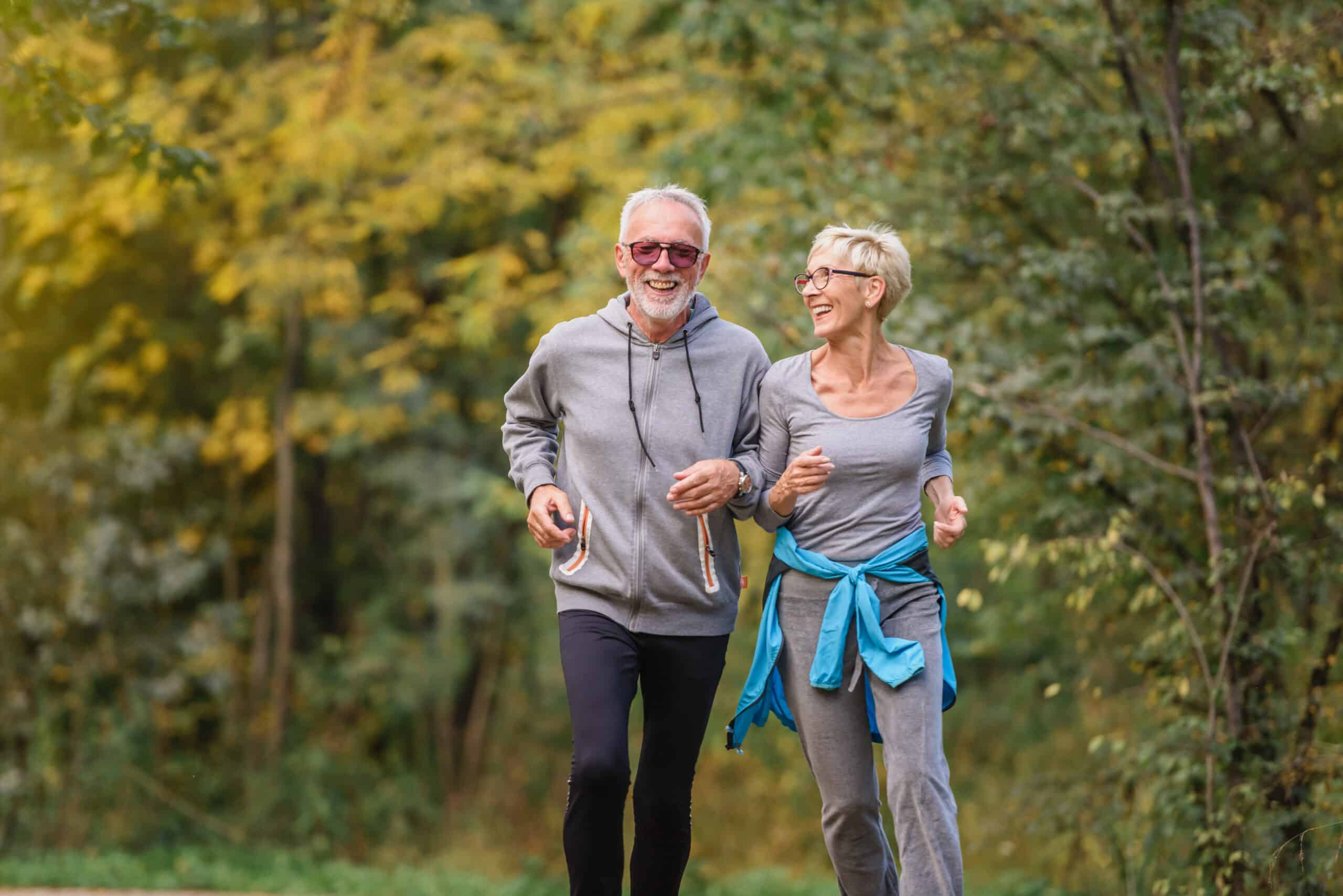 An older man and woman jog together outdoors on a wooded trail, smiling and wearing casual sportswear. Sunlight filters through green trees in the background.
