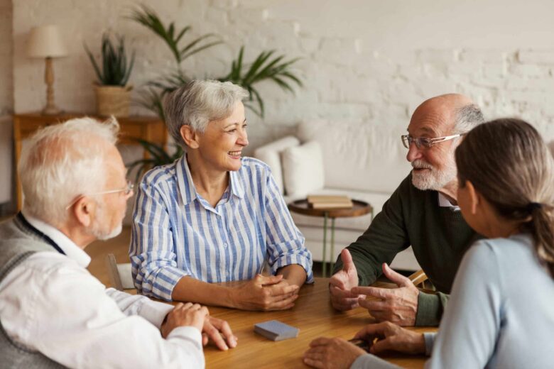 Four older adults sit around a table, smiling and talking together in a cozy room with plants and soft lighting. A deck of cards is on the table, highlighting the value of social connections as key Senior Care resources.