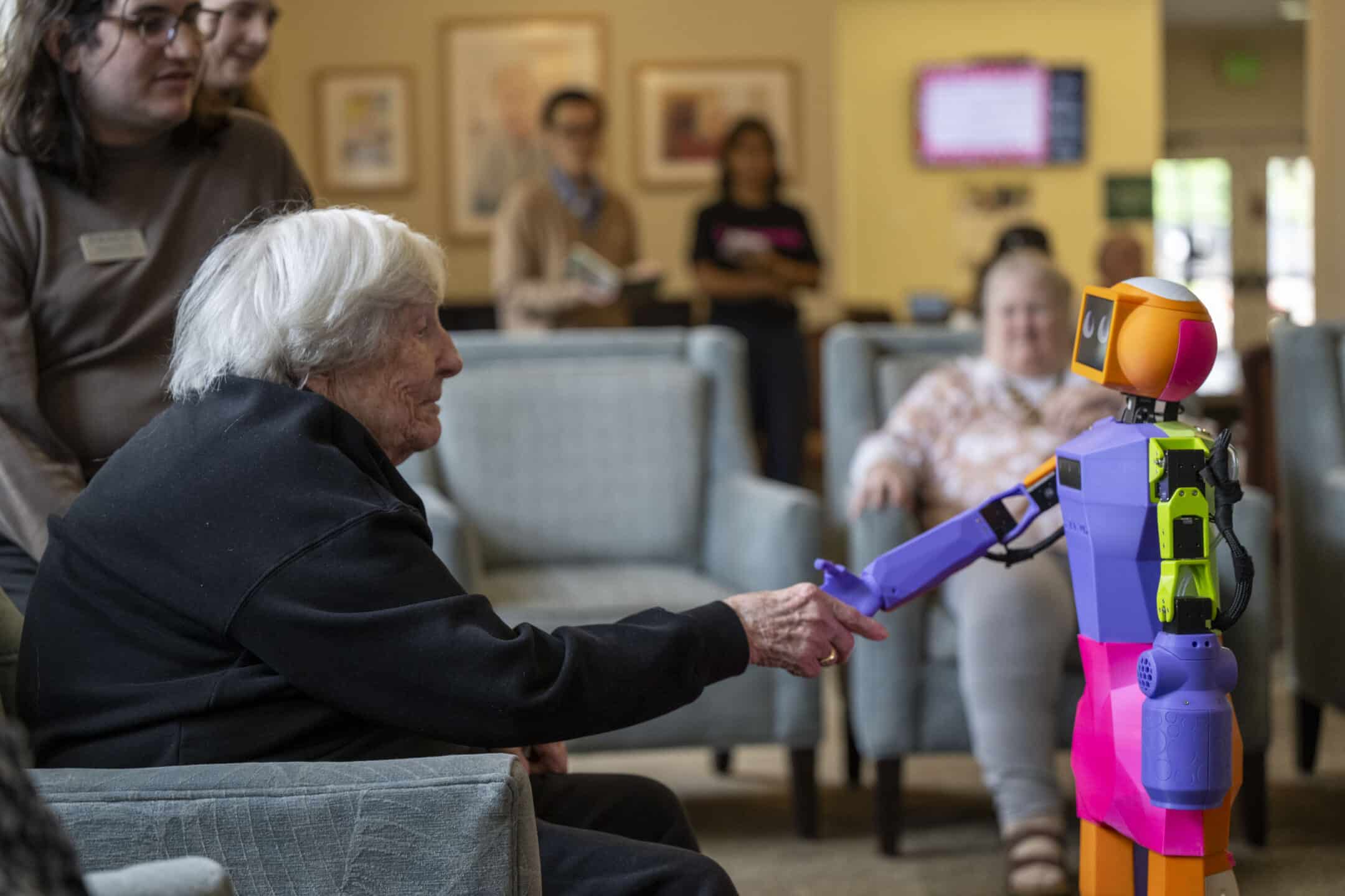 An elderly woman sitting in a chair shakes hands with a colorful humanoid robot, while several people watch and smile in the background in a bright, comfortable room.