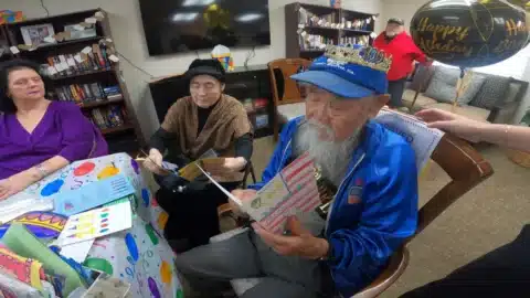 An elderly man wearing a birthday crown reads a card at a decorated table, surrounded by friends. Birthday balloons and cards are on the table, with people smiling and celebrating in a cozy room.