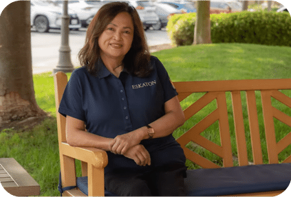A woman with medium-length brown hair, wearing a navy blue Eskaton collared shirt, sits smiling on a wooden bench in a park-like area with trees, grass, and parked cars in the background.
