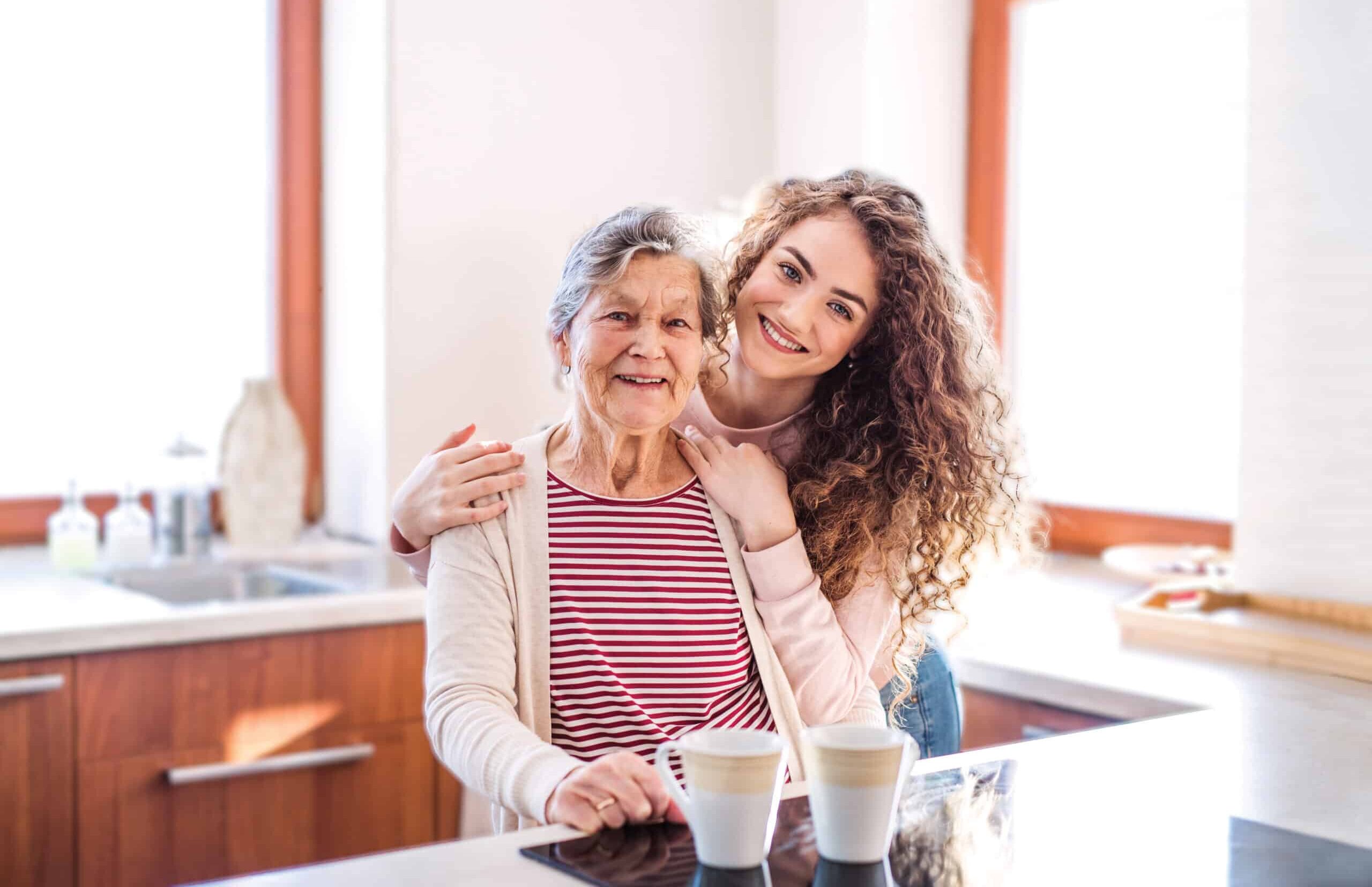 A smiling older woman and a young woman with curly hair pose together in a bright kitchen during a Community Spotlight Event, each with a cup of coffee on the counter. Large windows fill the space with natural light.