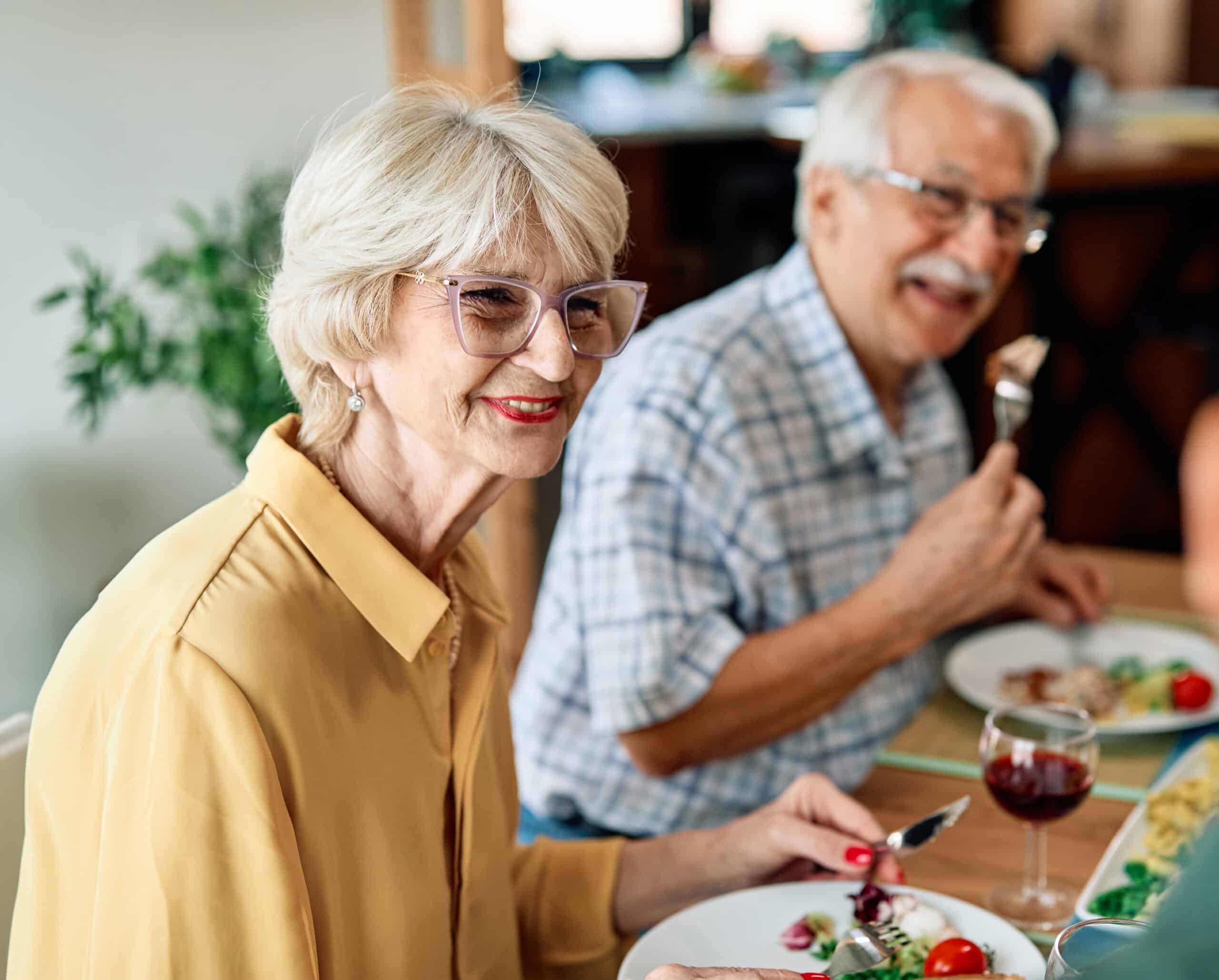 An elderly woman and man sit smiling at a dining table, enjoying a meal together. The woman wears glasses and a yellow blouse, while the man wears a plaid shirt and holds a fork. A glass of red wine and salad are visible.