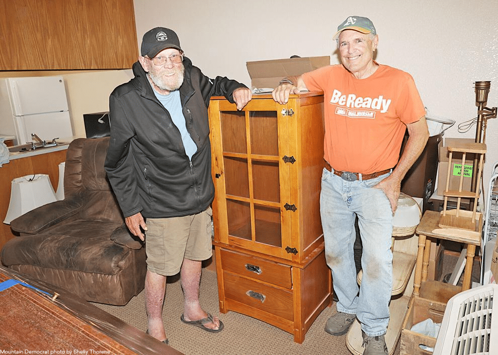Two older men stand smiling beside a wooden display cabinet in a room filled with furniture and household items. One wears a black jacket and cap, the other an orange Be Ready shirt and green cap.
