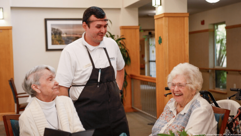 A man wearing an apron and face shield stands smiling with two elderly women seated at a table in a warmly lit common area, all appearing to enjoy a cheerful conversation.