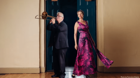 A man in a black suit plays a trombone beside a woman in a flowing, sleeveless, magenta floral gown. They stand indoors on a wooden floor with teal double doors and tan walls behind them.