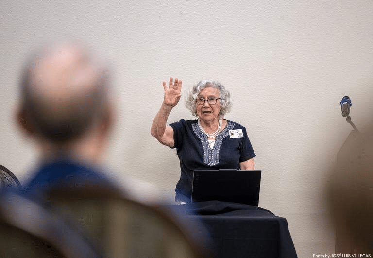 An older woman with gray hair and glasses sits at a table, raising her hand while speaking to an audience. A laptop is in front of her, and two people are visible in the foreground, facing her.