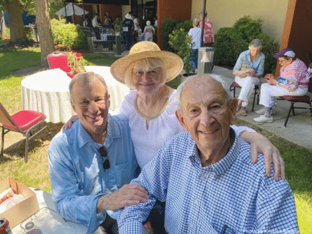 Three smiling seniors pose together outdoors at a gathering. The man on the left wears sunglasses, the woman in the middle wears a straw hat, and the man on the right sits wearing a checkered shirt. People relax in the background.