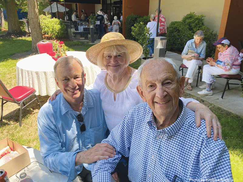 Three smiling seniors pose together outdoors at a gathering. The man on the left wears sunglasses, the woman in the middle wears a straw hat, and the man on the right sits wearing a checkered shirt. People relax in the background.
