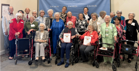A group of seniors, some in wheelchairs, pose together in front of an Eskaton banner. Most are smiling, and two people in the front hold certificates. Several people are standing behind them.