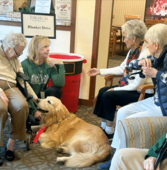 A golden retriever lies on the floor surrounded by four elderly women and a caregiver at a blanket drive event in a cozy room, creating a warm, friendly atmosphere.