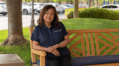 A woman with shoulder-length brown hair, wearing a navy blue shirt with the word ESKATON on it, sits and smiles on a wooden bench in a park-like outdoor setting with trees, grass, and parked cars in the background.
