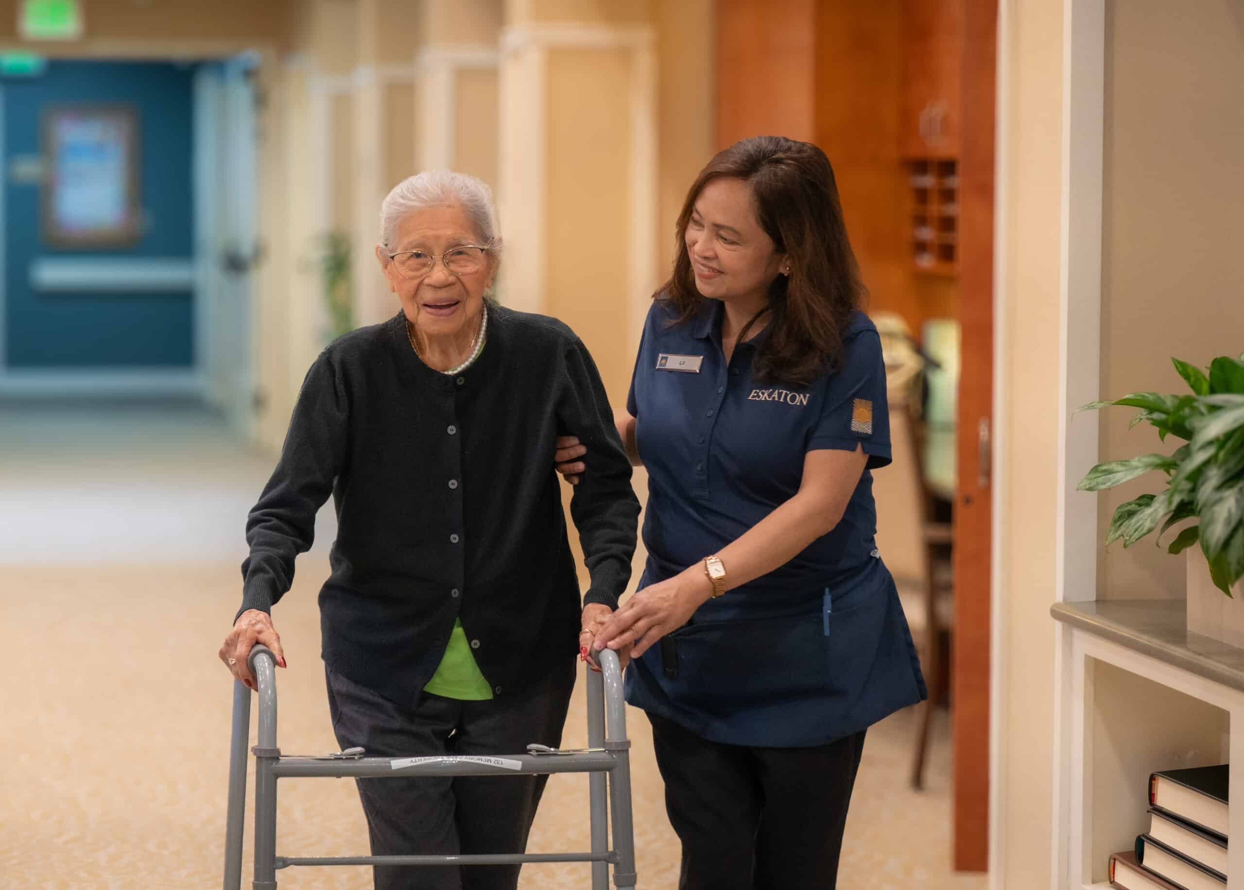 An elderly woman using a walker is assisted by a smiling caregiver in a hallway of a care facility. The caregiver gently supports the woman as they walk together.