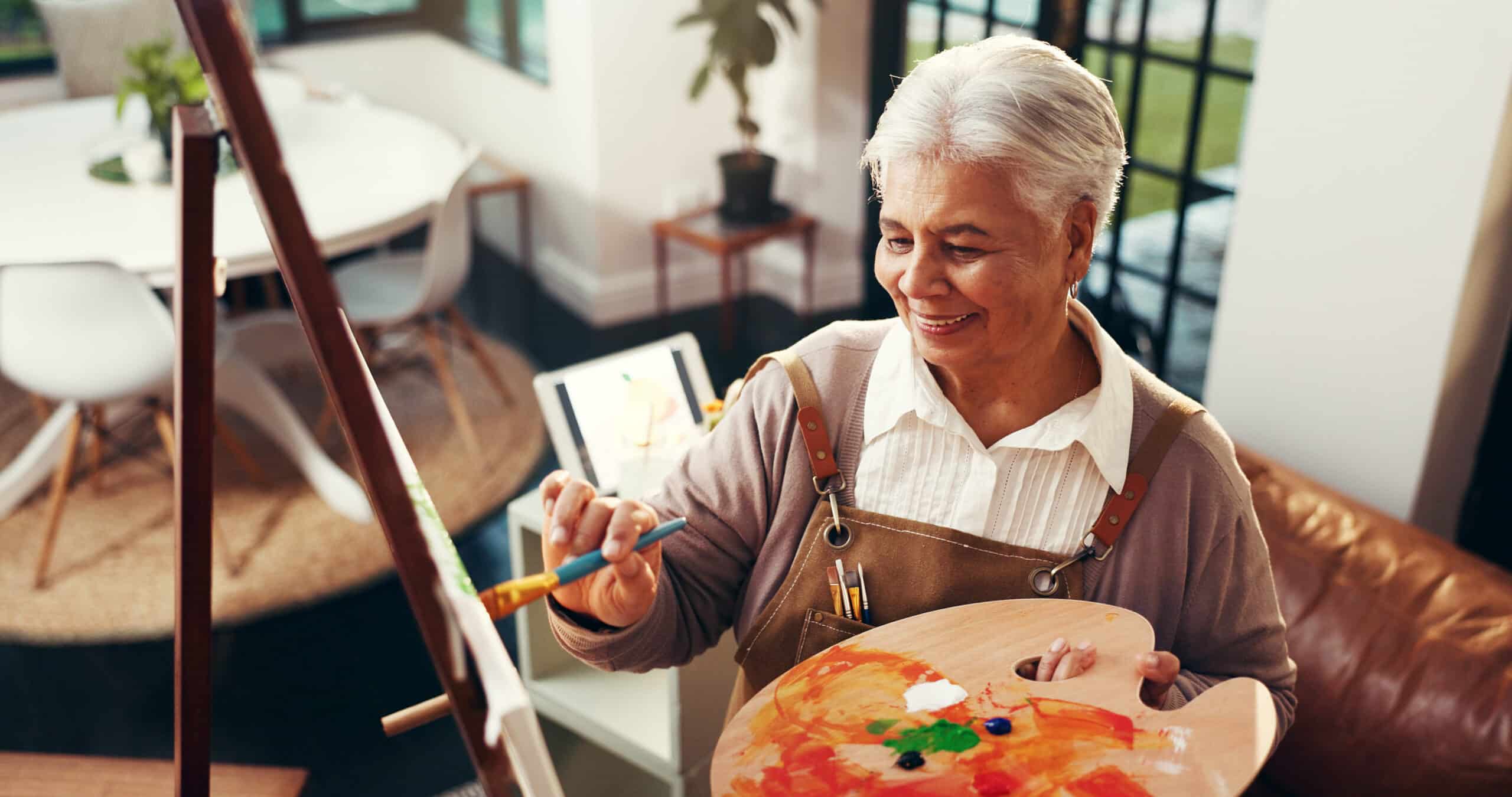 An older woman with gray hair smiles while painting on a canvas indoors, holding a palette with colorful paint. She wears an apron and stands in a bright, modern room with large windows and plants.