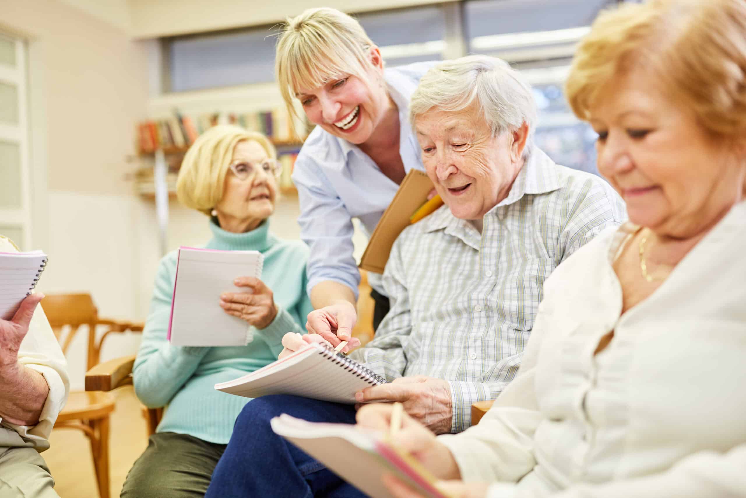 A group of older adults sit together in a classroom, holding notebooks and smiling, while a woman stands and helps one man with his writing. Bookshelves are visible in the background.