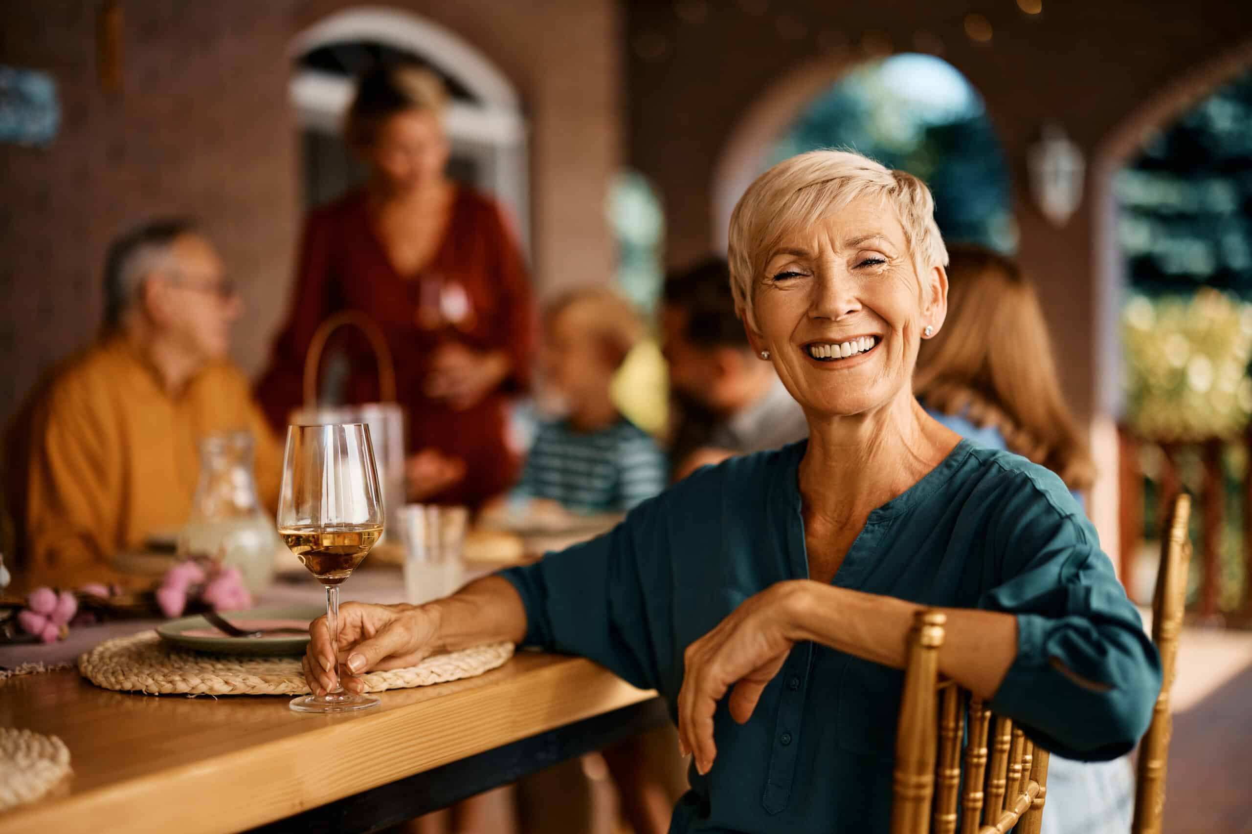 Smiling older woman holding a glass of wine sits at an outdoor table, with family or friends gathered and talking in the background on a sunny day.