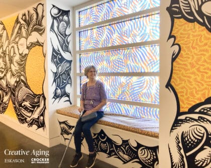 An older woman sits on a window bench in front of colorful, abstract-patterned glass and bold, black-and-white wall art. Creative Aging and museum logos appear in the bottom left corner.