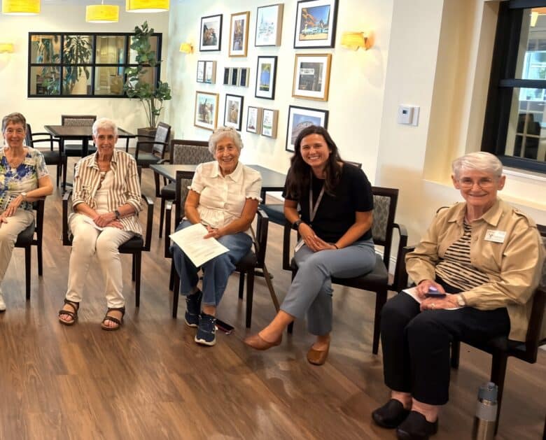 Five women sit in chairs in a brightly lit room with wooden floors, framed photos on the wall, and large windows. They are smiling and appear to be in a casual group meeting or gathering, fostering an atmosphere of innovation and collaboration.