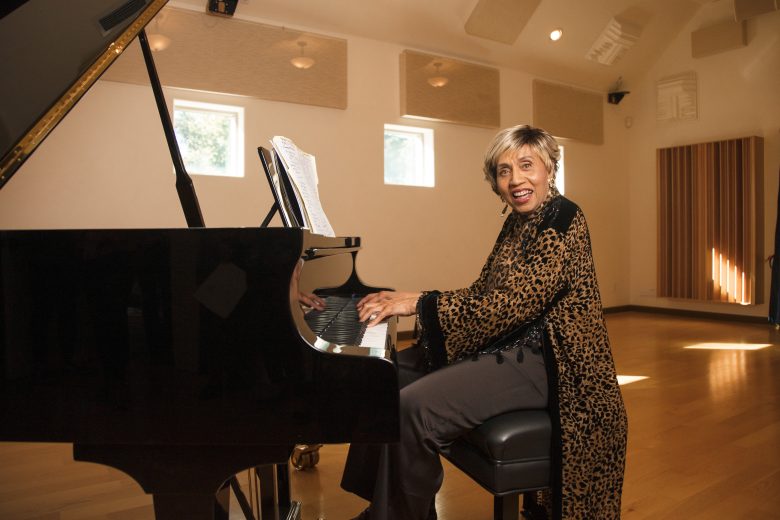 An older woman in a leopard-print outfit sits at a grand piano in a sunlit room, smiling toward the camera, with sheet music on the piano.
