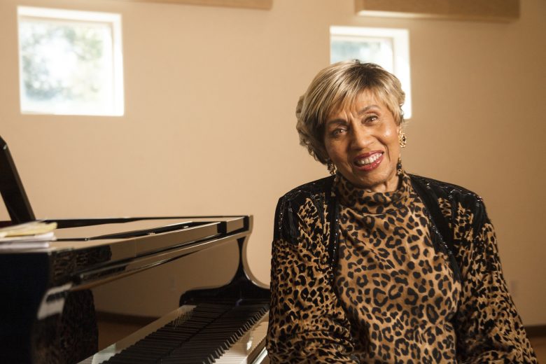 An older woman with short, light hair and a leopard print top sits and smiles beside a grand piano in a bright room with beige walls and three square windows, embodying a lifelong passion for music.