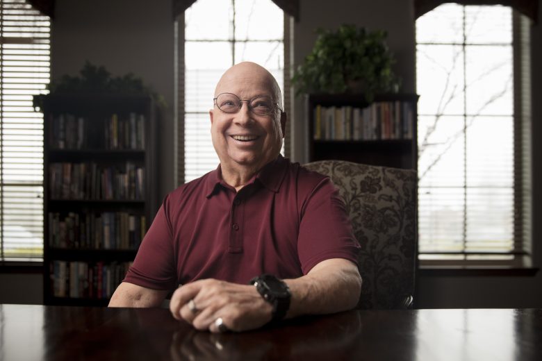Smiling older man wearing glasses and a maroon polo shirt sits at a dark table in a room with bookshelves, large windows, and a plant in the background.