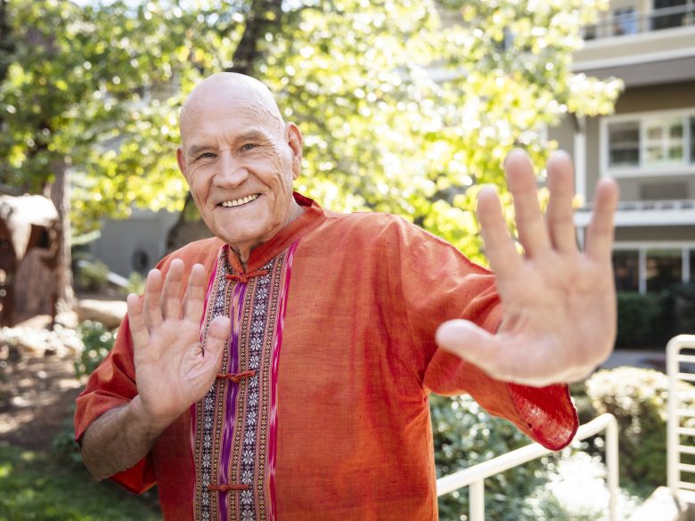 Smiling older man in an orange traditional shirt stands outdoors, raising both hands with palms facing forward, as if greeting or gesturing in a welcoming manner. Sunlight and greenery are visible in the background.