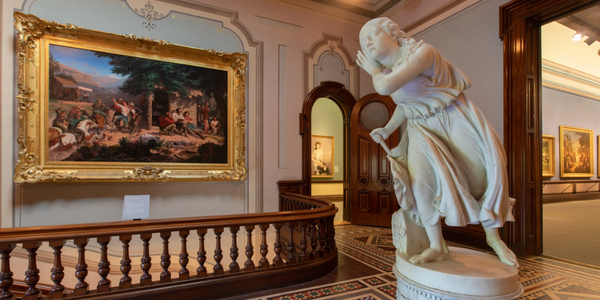 A marble statue of a woman stands near a wooden railing in an ornate gallery, with framed paintings on the walls and intricate floor tiles visible.