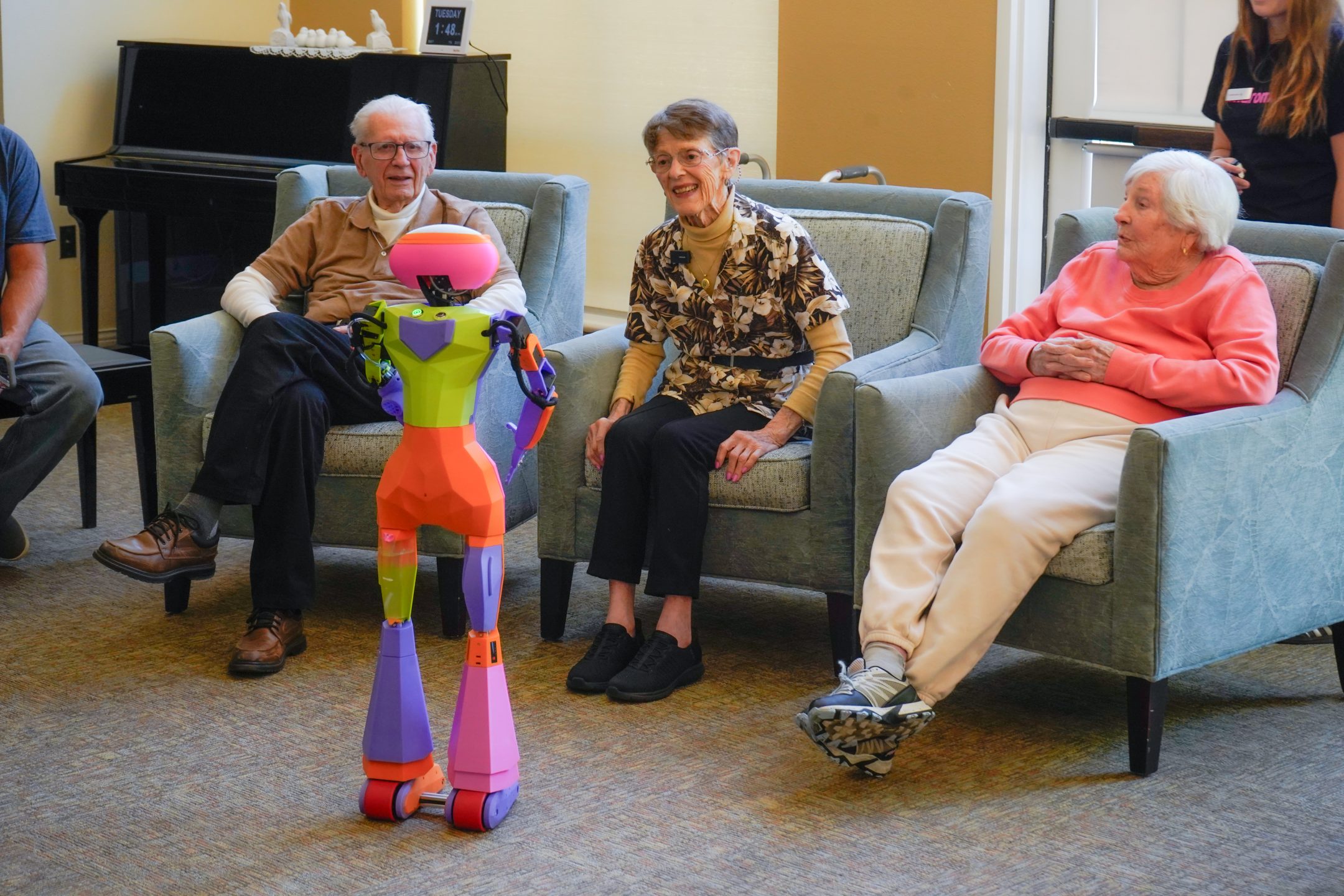 Three elderly people sit in armchairs, smiling and watching a colorful robot standing in front of them. A piano is visible in the background, and someone stands near a door on the right.
