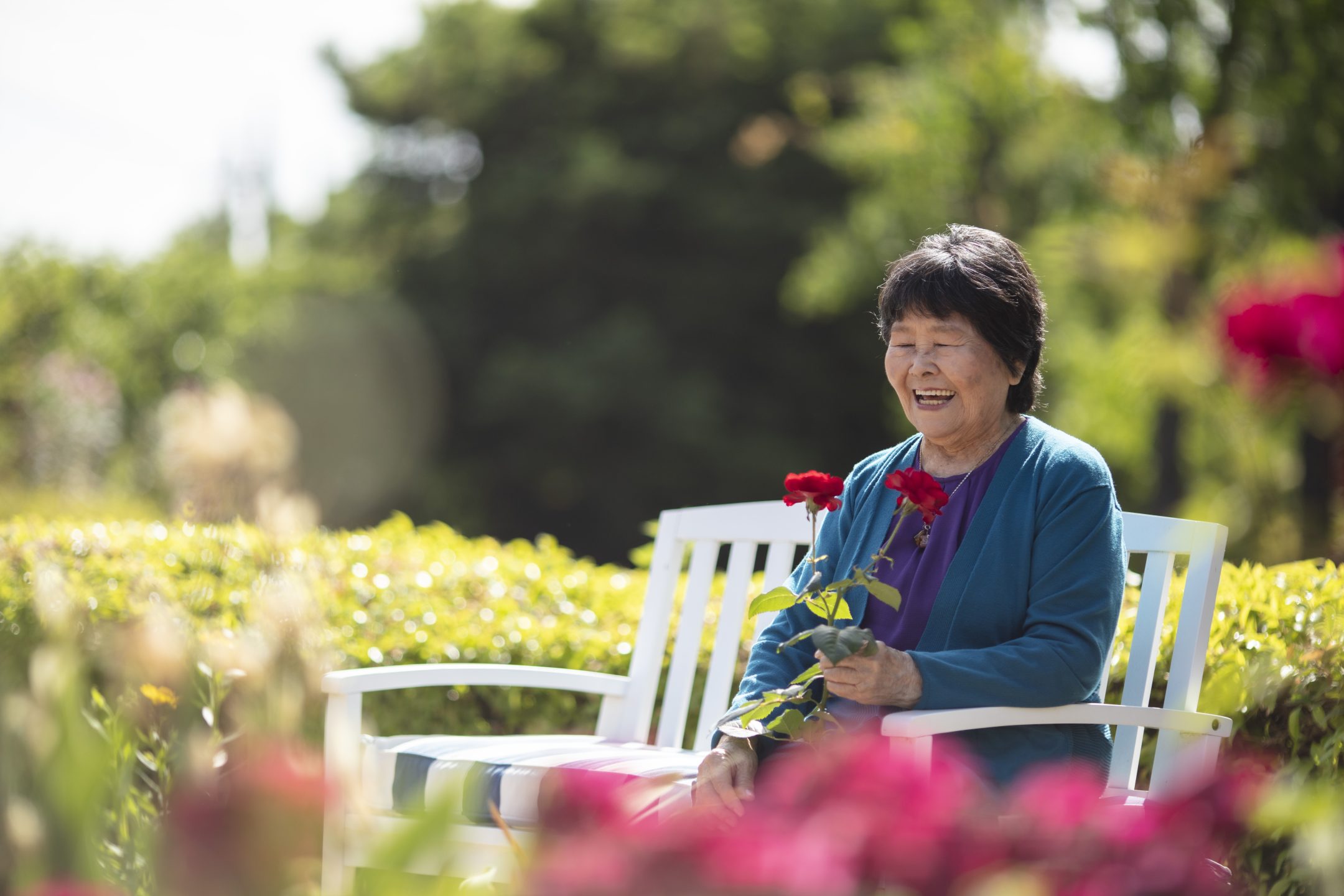Elderly woman sits on a white bench in a garden, smiling and holding red roses, surrounded by greenery and flowers on a sunny day.