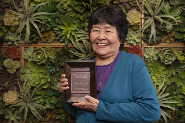 Smiling woman in a blue cardigan holds a Volunteer Spirit Award plaque. She stands in front of a wall decorated with green and succulent plants.