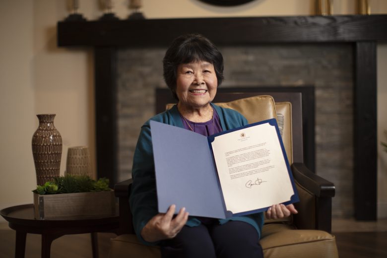 Smiling elderly woman sits in an armchair holding an open folder with a formal letter inside, perhaps recounting a cherished tai chi tale, in a warmly decorated room with vases and plants.