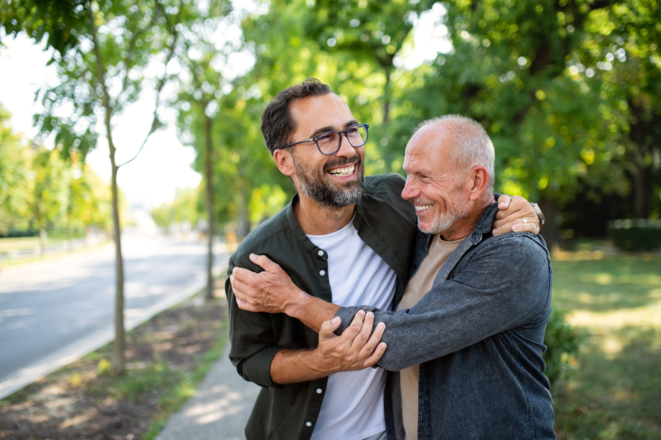Two men smiling and hugging each other while standing on a tree-lined sidewalk on a sunny day.