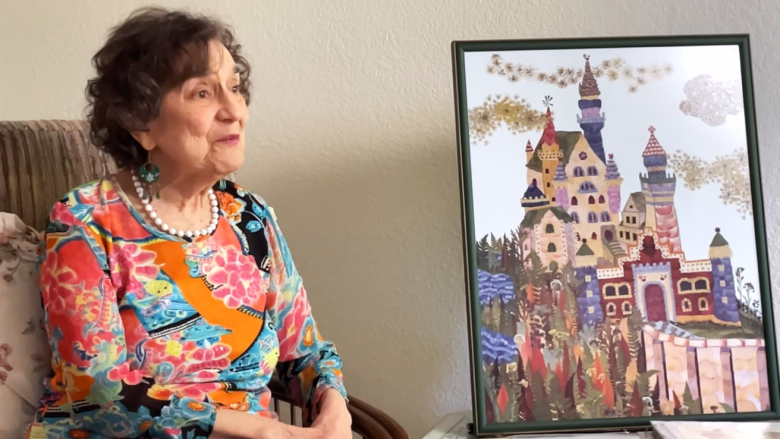 An older woman in a colorful dress and pearl necklace sits in a chair next to a framed painting of a whimsical castle. The background is a neutral indoor wall.
