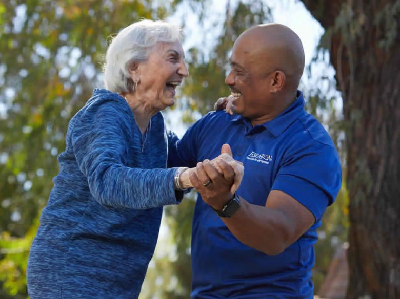 An elderly woman and a man smile and hold hands while dancing outdoors, both appearing happy. The man wears a blue shirt, and trees fill the background.